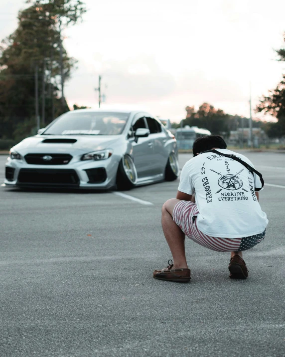 a young man squatting down in a parking lot with his backpack on his back and a silver car behind him