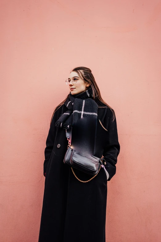 a woman is standing near a pink wall wearing glasses and a trench coat