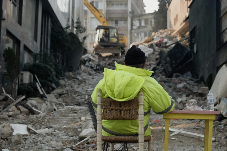 a person is sitting on the chair next to a broken table and a large building with a bulldozer in it