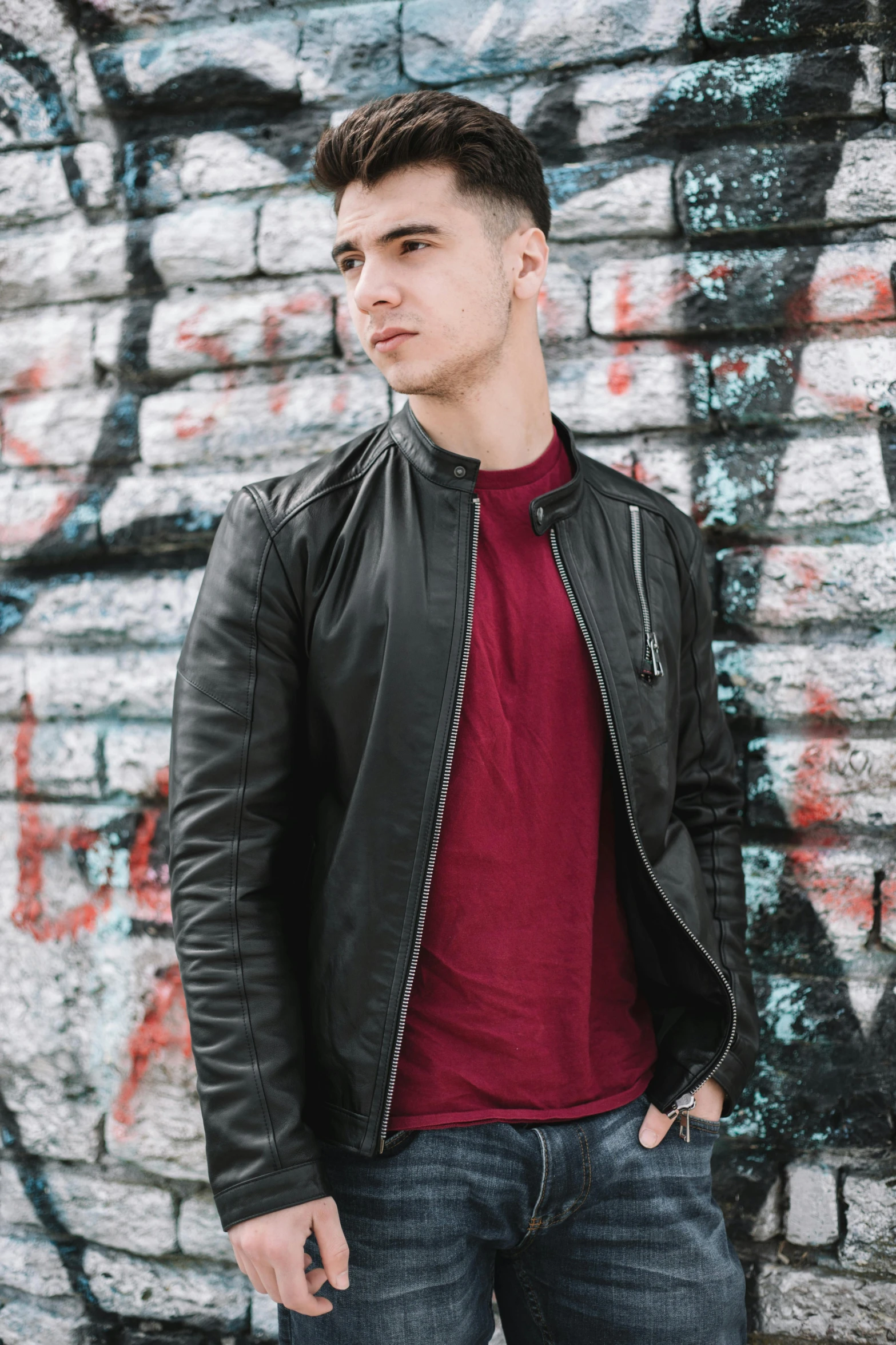 a young man in a red shirt and black leather jacket standing by a brick wall