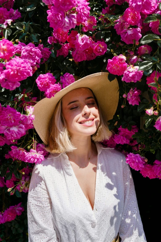 a woman wearing a sun hat smiling near a flowered bush