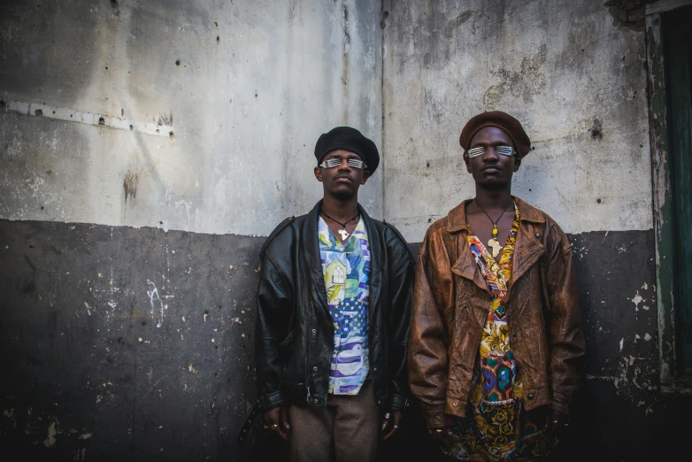 two black men stand by a wall wearing glasses and jackets