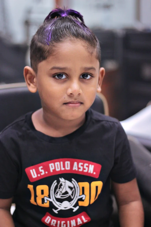 a young black child wearing a u s polo assn t shirt with an orange and black stripe on the front of the shirt, sitting in front of an office chair