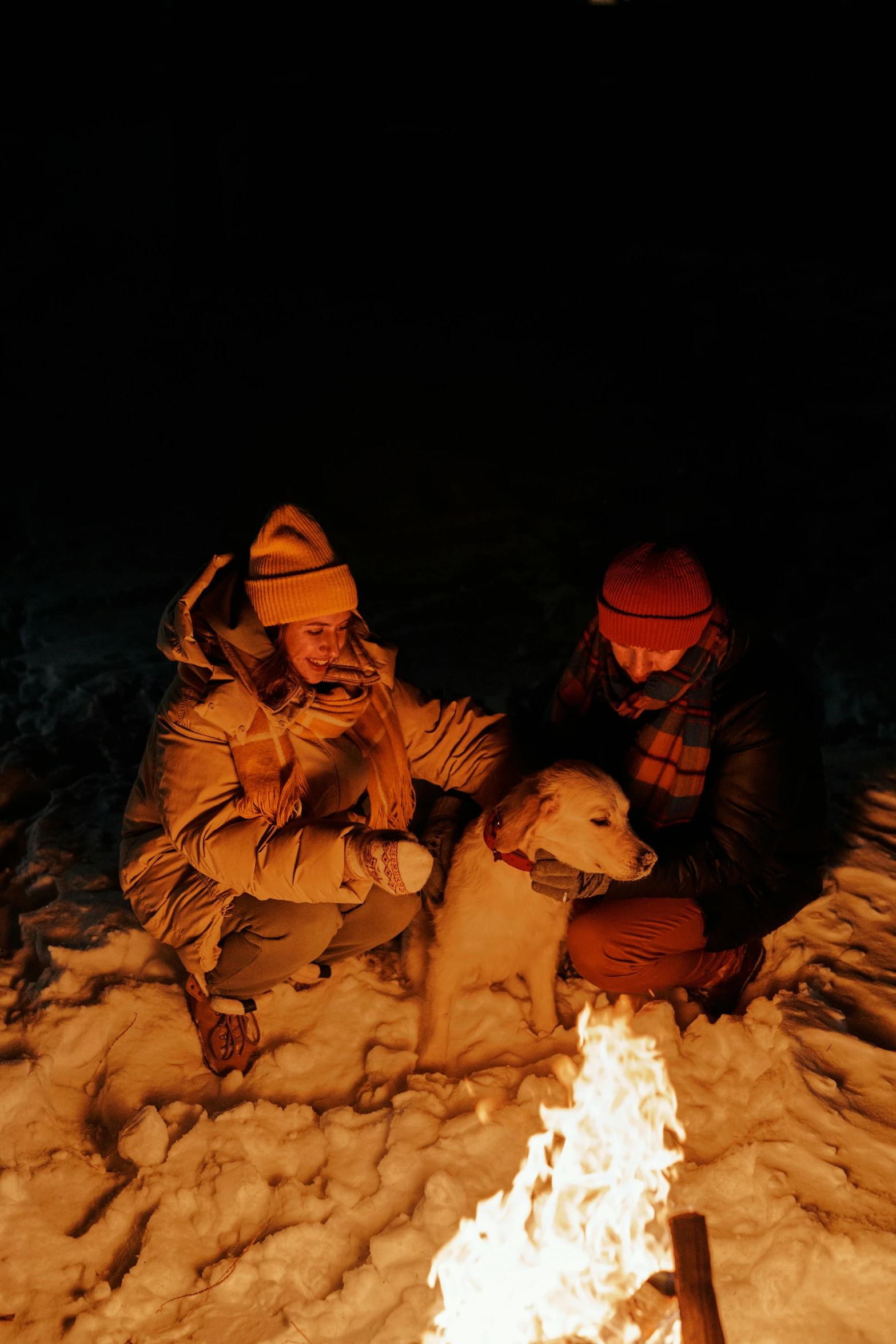 a couple sit in the snow with their dog next to a campfire