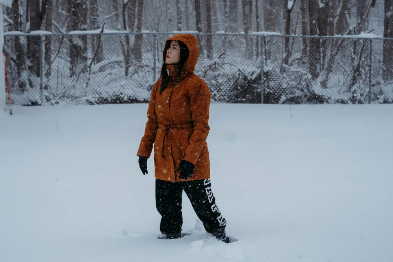 woman in orange jacket standing in snow in front of a fence