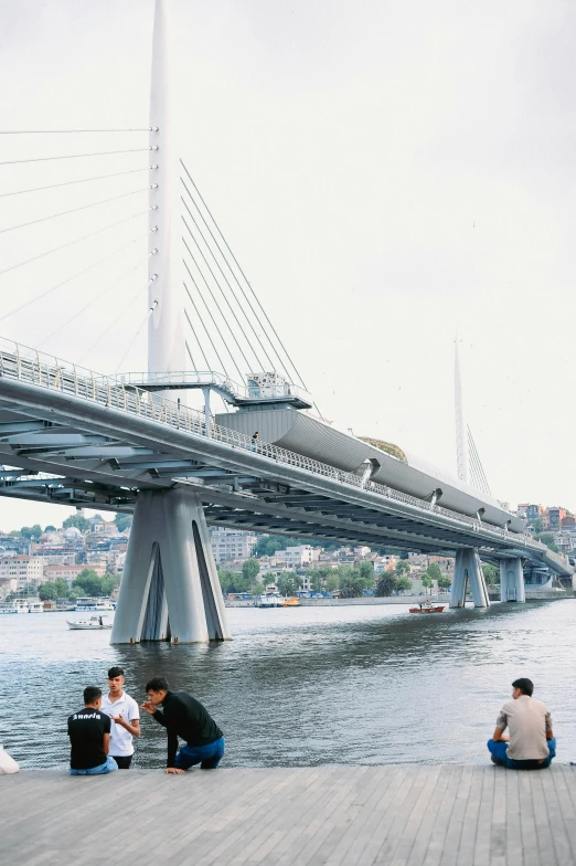 three people sitting in the sun on a deck looking at a bridge over water