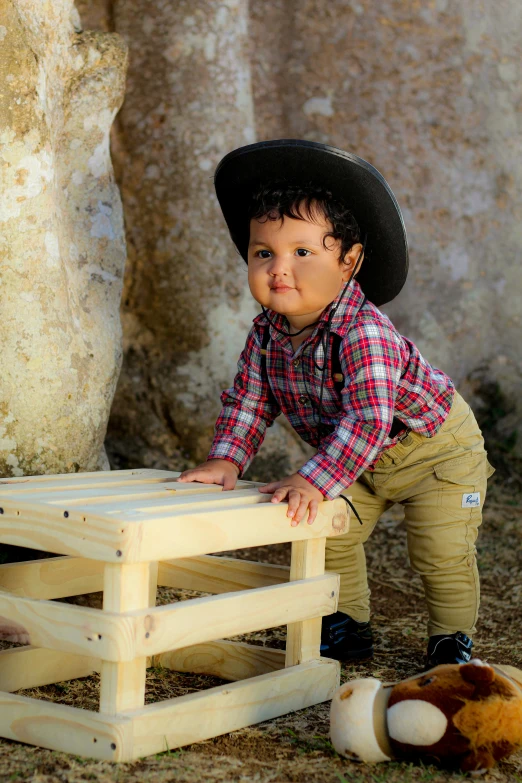 a boy wearing a cowboy hat, shirt and green pants sitting in a wooden pallet with a toy horse