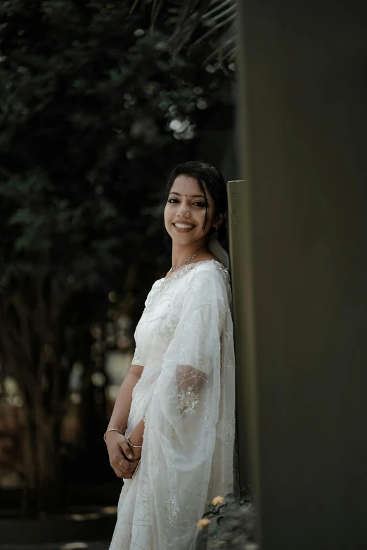 a smiling woman in a white sari stands in front of a tree