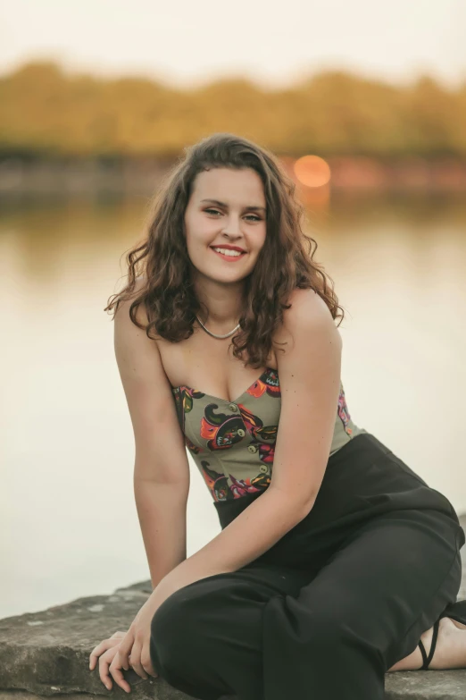 a woman in a floral shirt and black pants sits on rocks near a lake