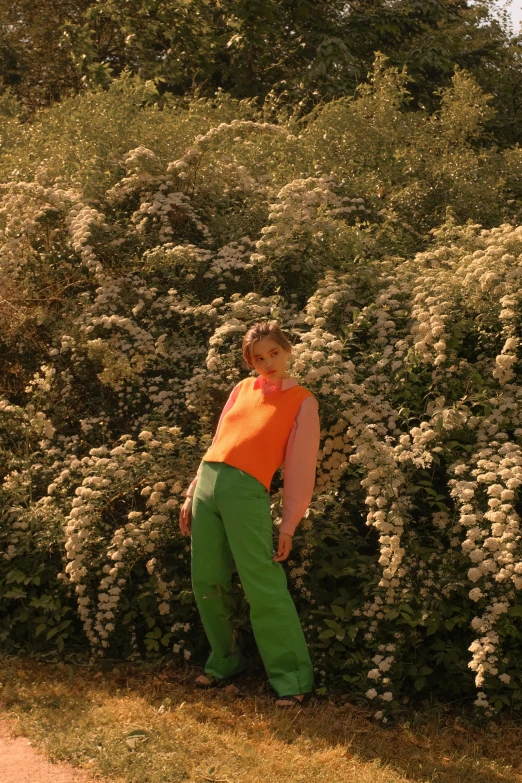 a  wearing a orange shirt and green pants stands by a flower bush