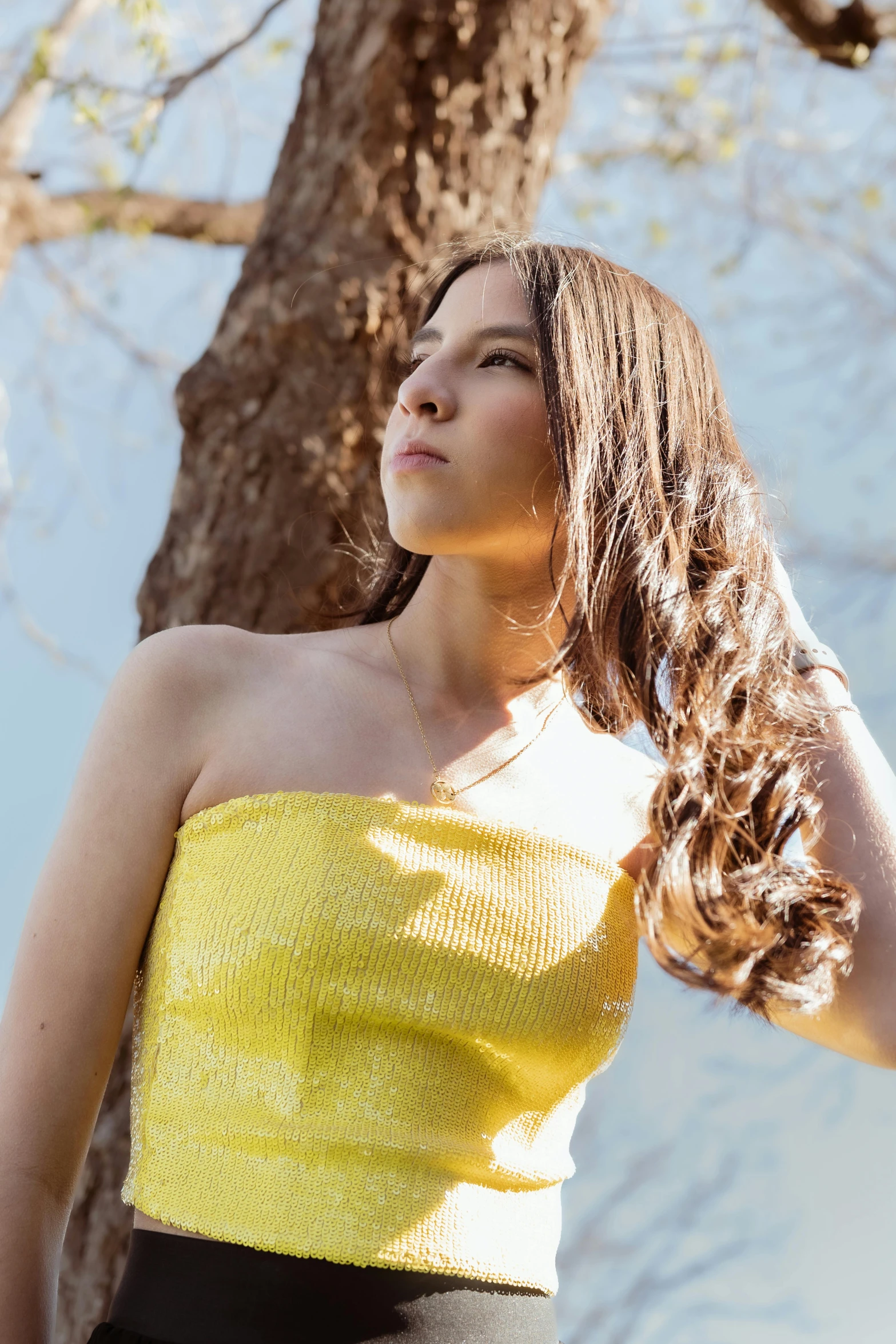a woman with brown hair, in front of a tree, is wearing a yellow top