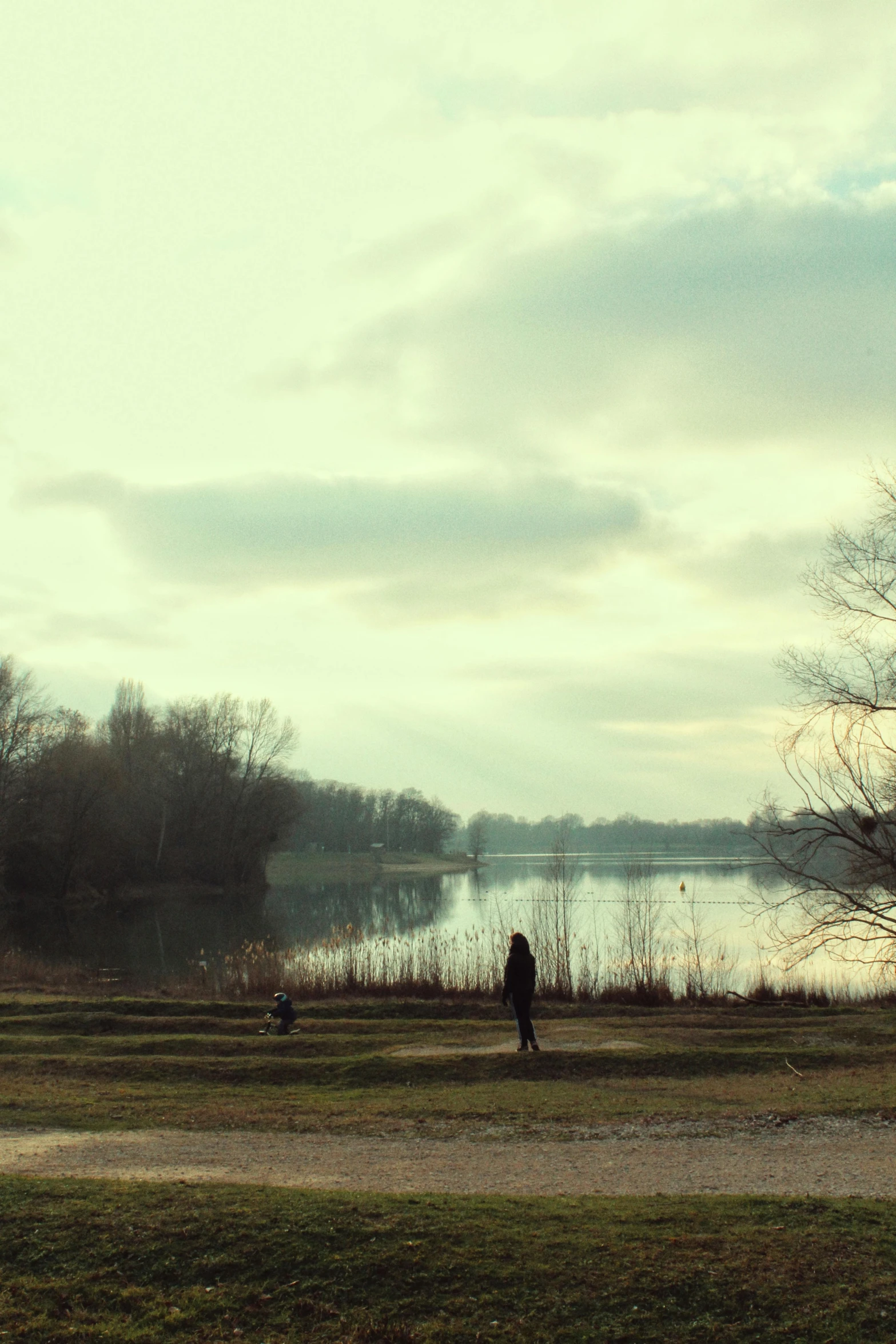 two people sitting and standing on a grassy field next to the water