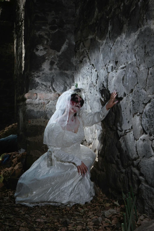 the woman in white and wearing makeup sits against a wall