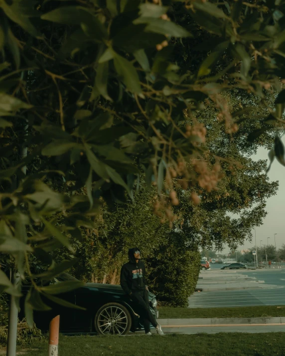 a man in black jacket and jeans sitting on top of a car near trees