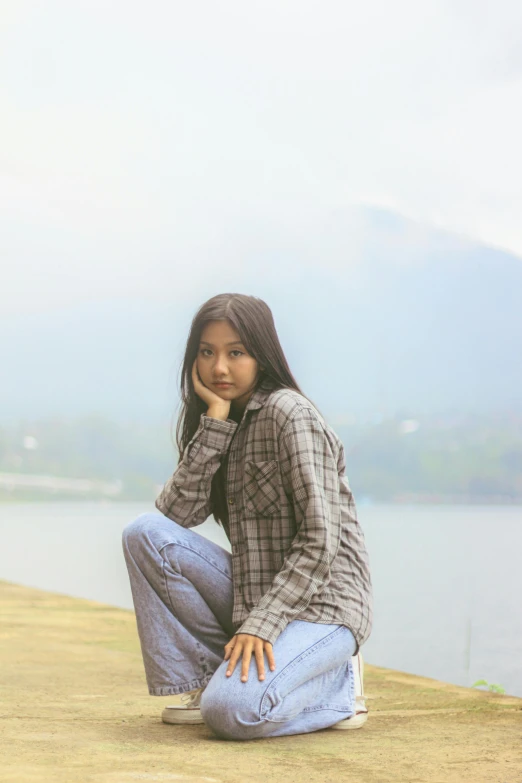 young woman with long dark hair, in jeans sitting on a dock by a lake