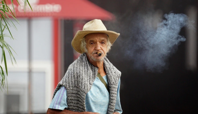 an old man wearing a hat, coat and jacket smoking a cigarette