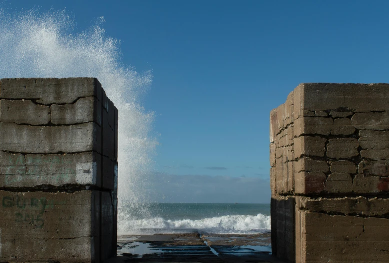 two gray blocks are facing the water with white foam crashing from the rocks