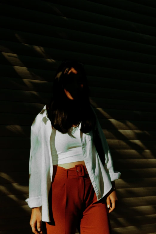 the woman stands against a wall as she wears a white top and maroon pants