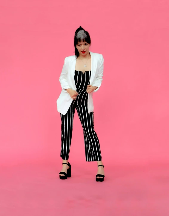 a  in a white cardigan and striped pants stands against a pink background