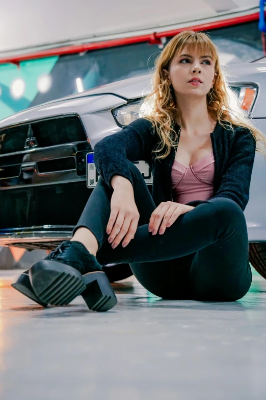 a woman in a pink shirt and black jacket sitting on the ground near a car