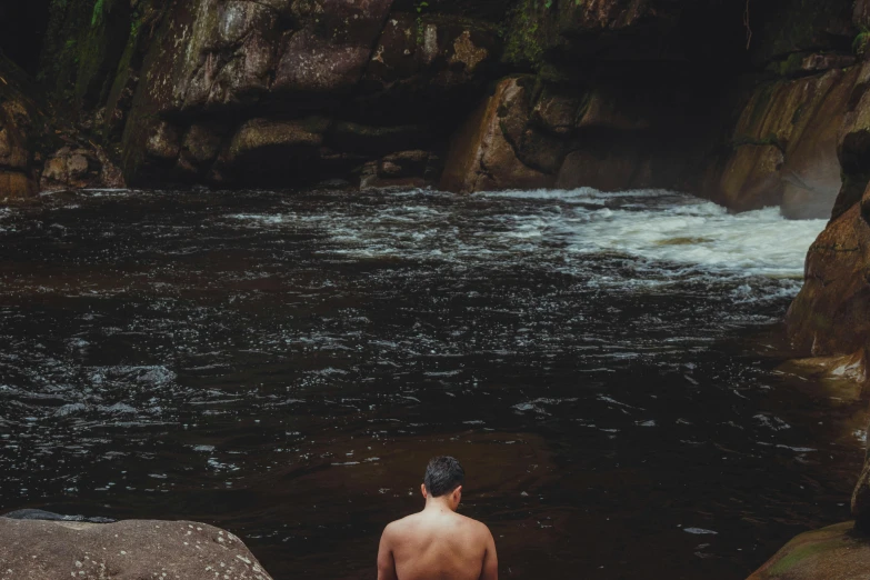 a man in a black swim suit standing in a river by some rocks