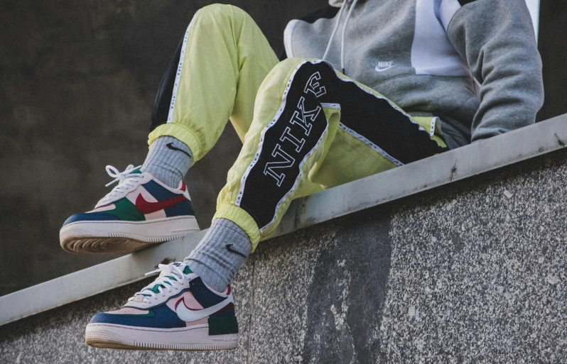 a person wearing green pants, gray sweater and colorful sneakers sits on a wall