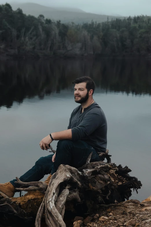 a bearded man sits on a tree stump next to a river