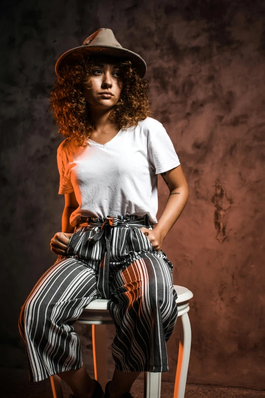 a girl wearing a hat and a striped pants sits on a stool