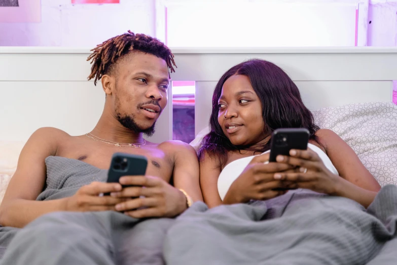 two people lie on bed looking at their cellphones