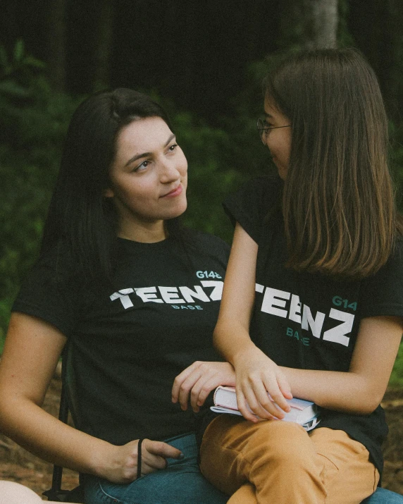 two women wearing t - shirts sitting next to each other in the woods