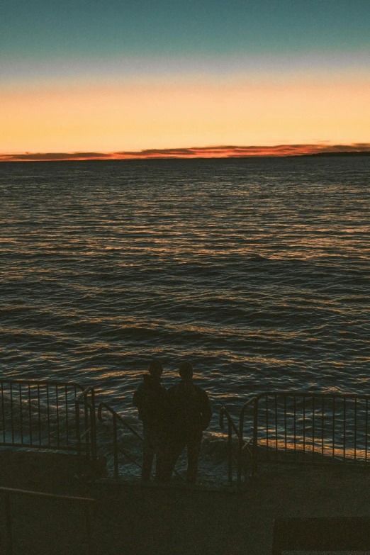 two people standing near the ocean with a setting sun