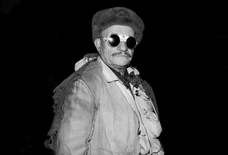 an older man in round glasses standing with a hat on