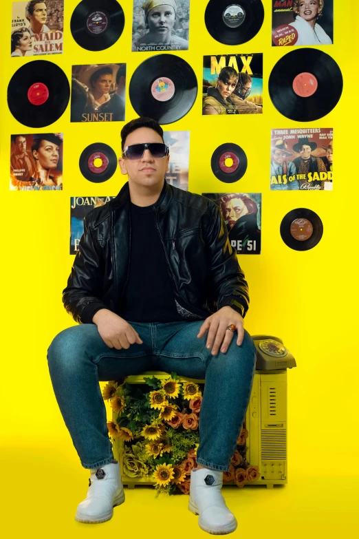 a man in a black leather jacket sits on a stool with his feet up against a wall covered with vinyl records