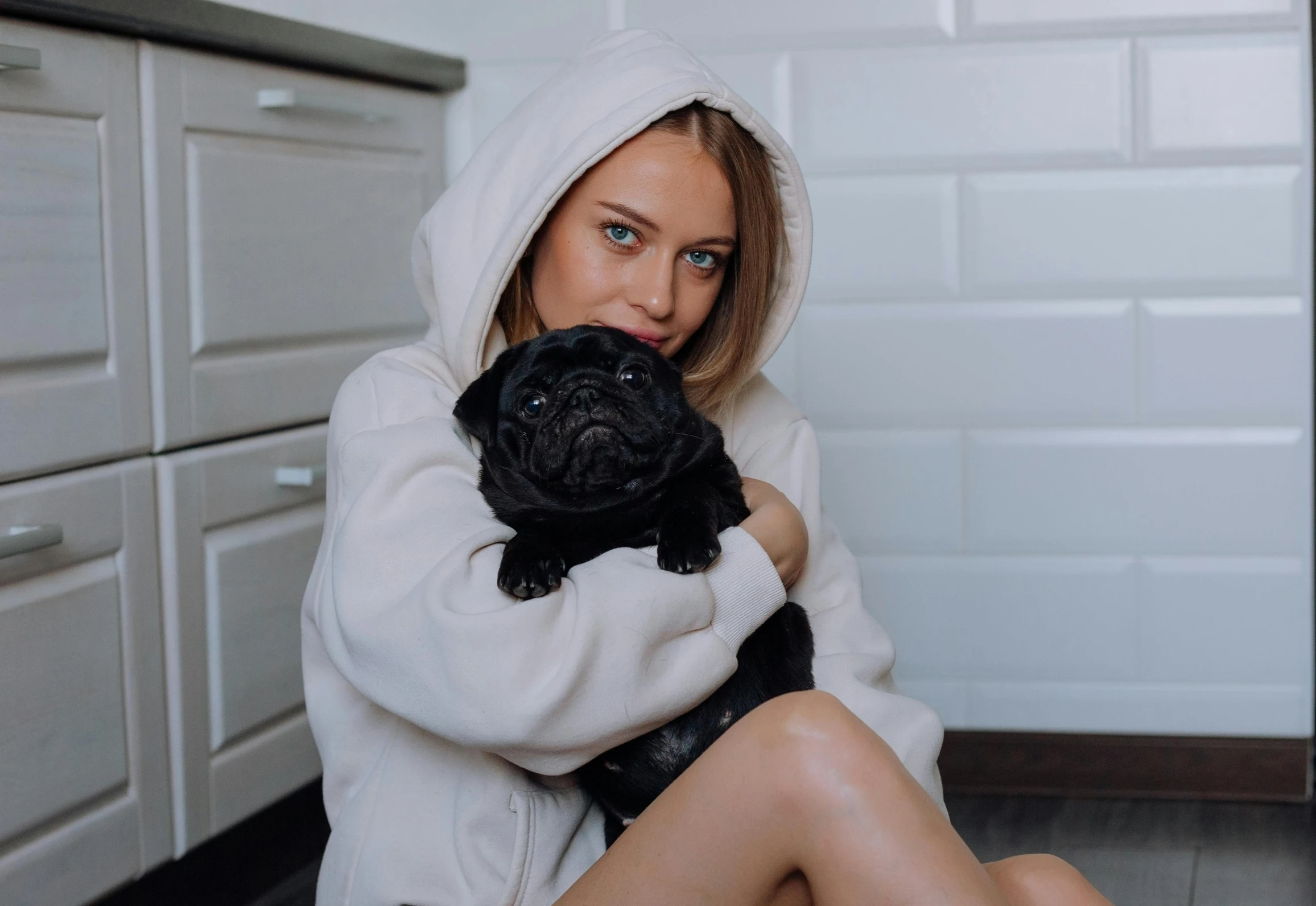 a woman wearing a white sweater holds her pug while sitting on the kitchen floor