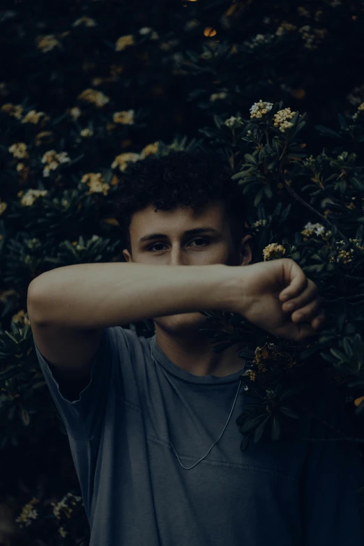 a young man in grey shirt leaning against bushes