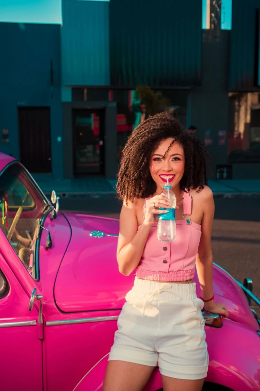 a girl smiling while drinking water next to a vintage car