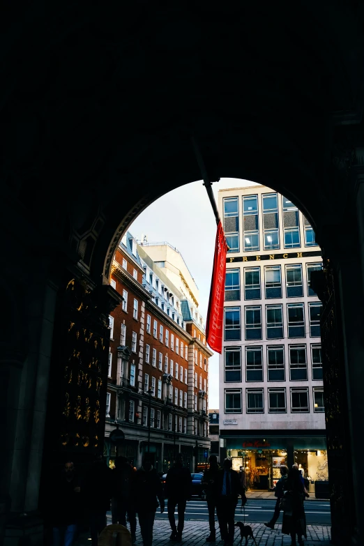 people walking in the shade at the corner with tall buildings and red flags