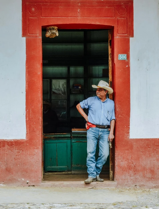 a man in a cowboy hat leans against a red building's front door