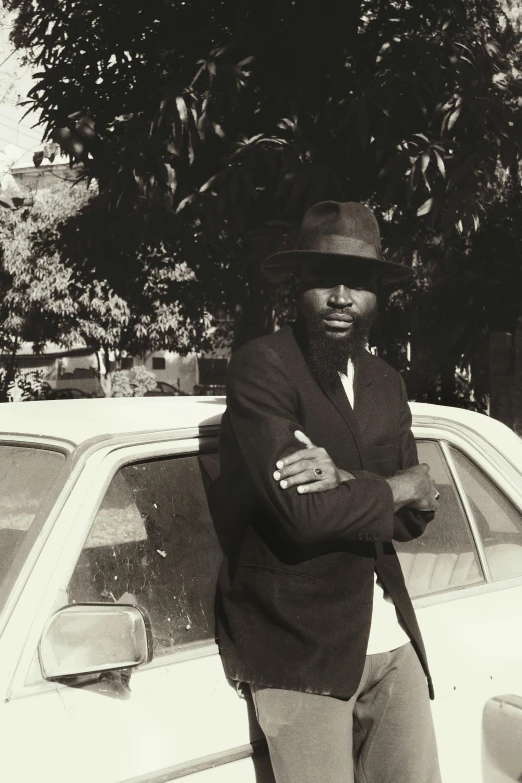 a man in hat, jacket and dress pants standing near a car