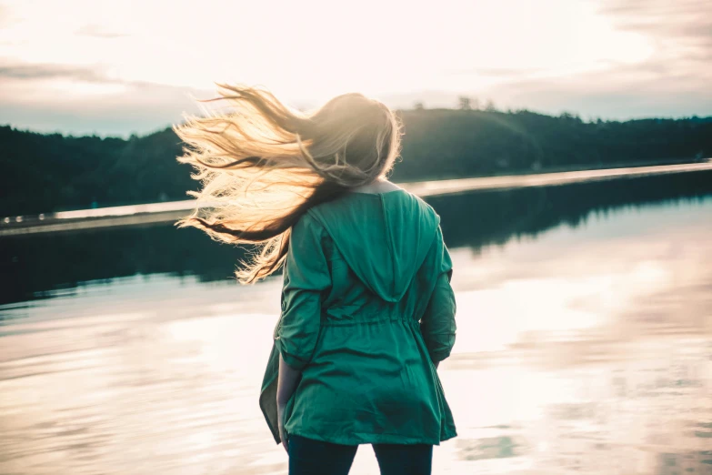 a woman stands on the shoreline with her hair blowing in the wind