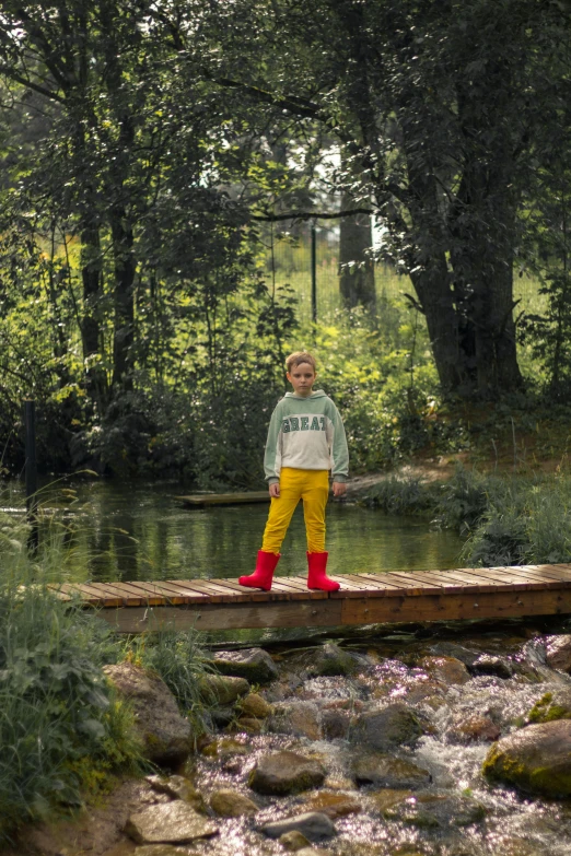 a boy in red boots and yellow pants stands on a bridge