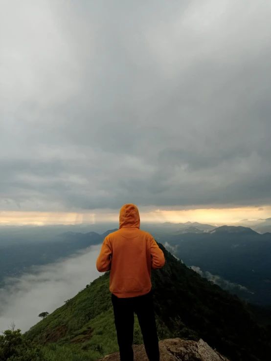 a person wearing an orange jacket and facing a hill under a cloudy sky