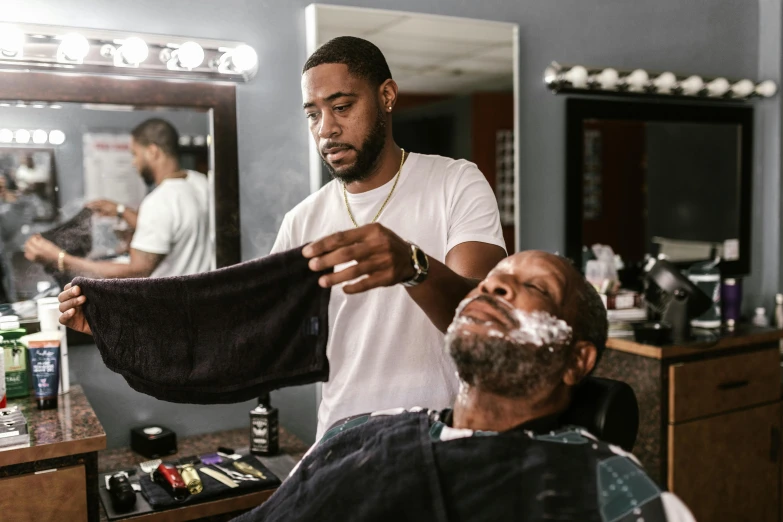 an old man shaving at the barber shop with another man in the chair nearby