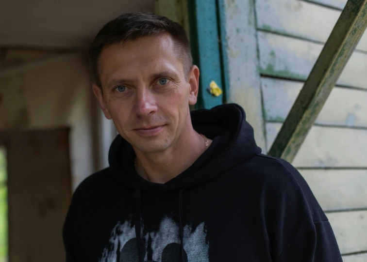 a young man wearing a dark shirt standing in front of a wooden house