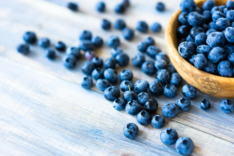 a wooden bowl filled with blueberries spilling out on a white wooden surface