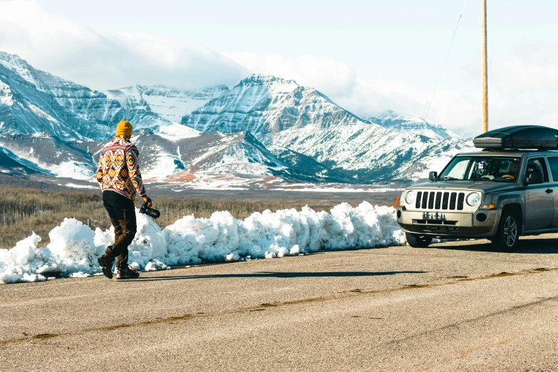 man walking on road with vehicle nearby with snow piled on mountains in the background