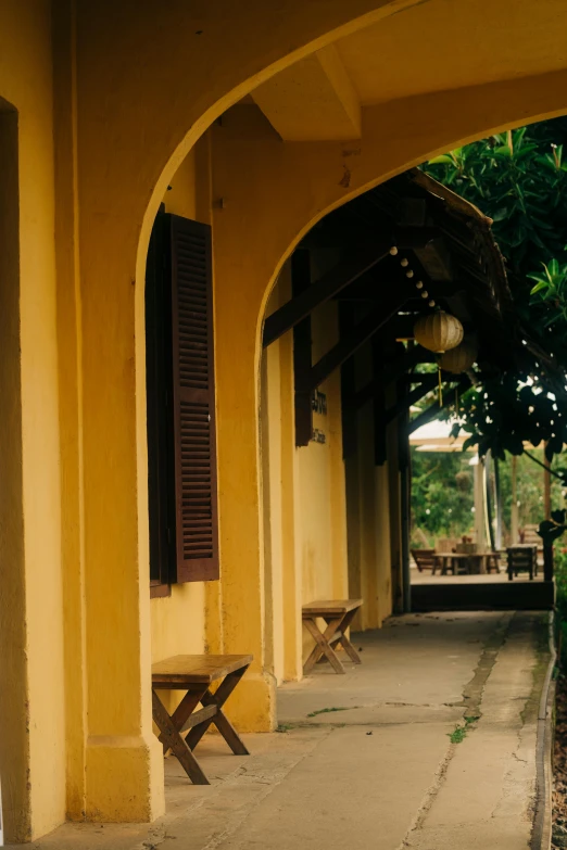 an empty pathway in front of a yellow building with three wooden benches under a roof