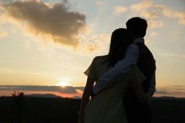 a man and woman standing on top of a hill in front of a sunset