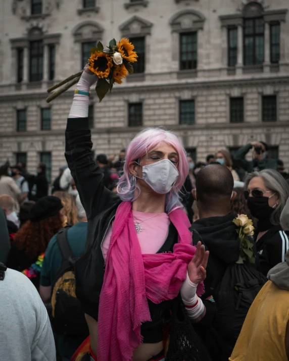 a young person with pink hair wears a mask and holds a flower