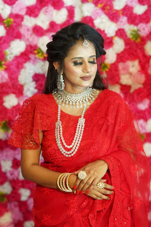 a woman with long hair in a red sari and gold jewelry stands next to a flower wall